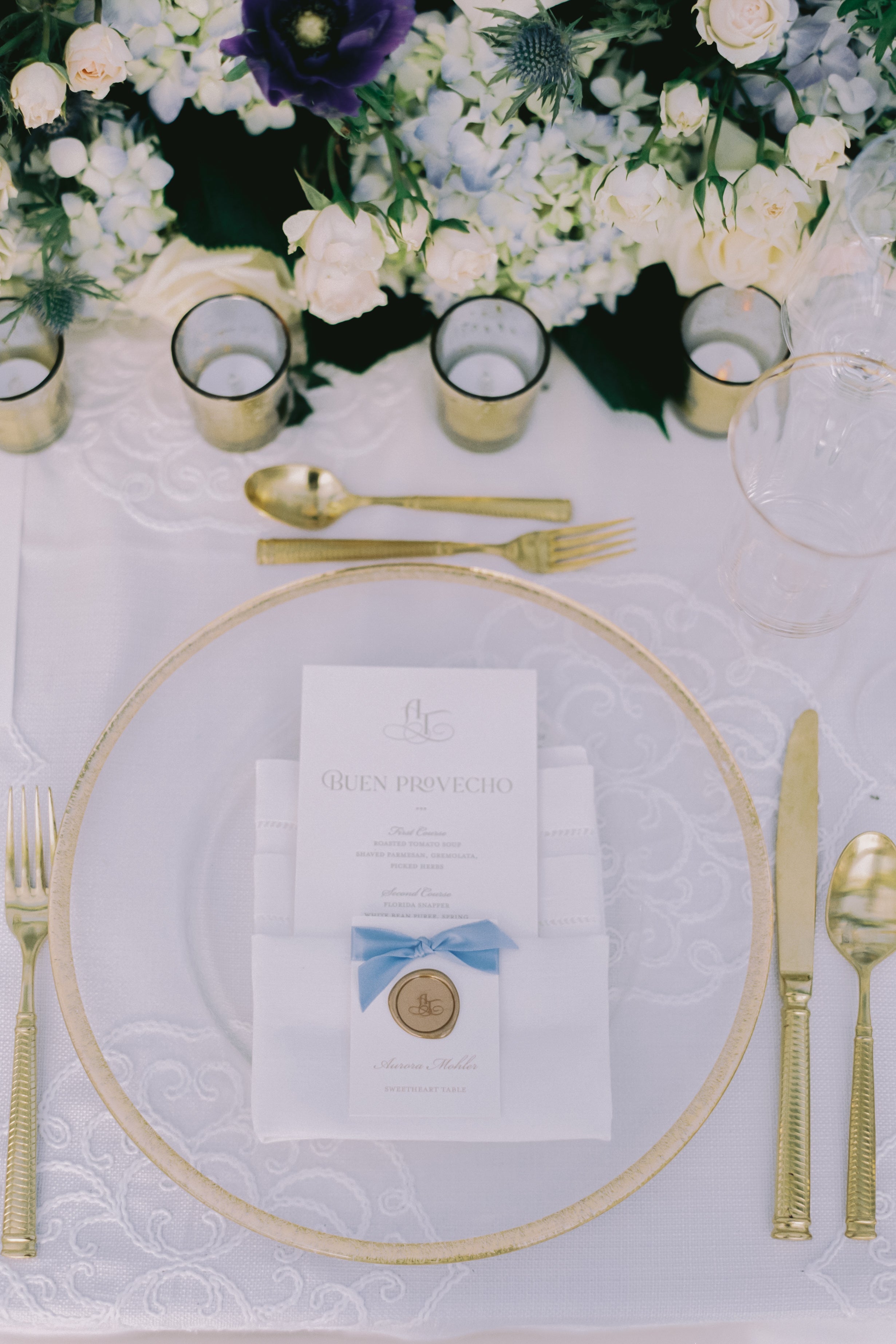 Elegant table setting with gold cutlery, white napkin with blue bow, and floral decorations.